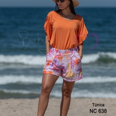 Mulher na praia com túnica laranja e calções florais, chapéu de palha e óculos escuros.