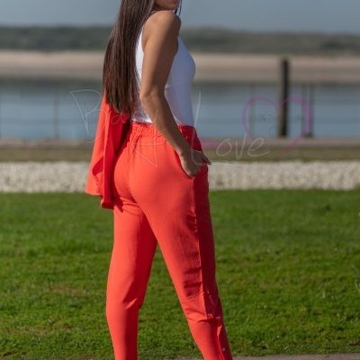 Mulher de perfil com calças laranja e top branco ao ar livre junto à água.