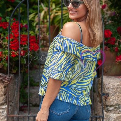 Mulher com t-shirt azul e amarela de folhos e jeans azuis, usando óculos escuros, em jardim com flores e cerca de ferro