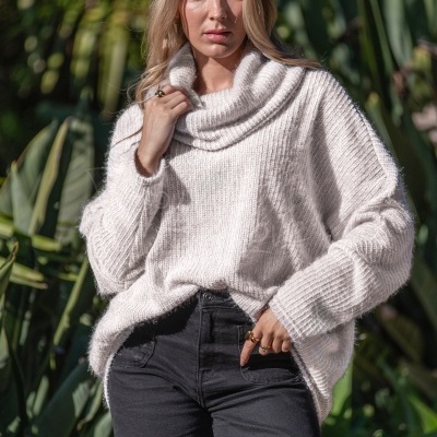 Mulher com camisola creme de malha e calças pretas em fundo de plantas.