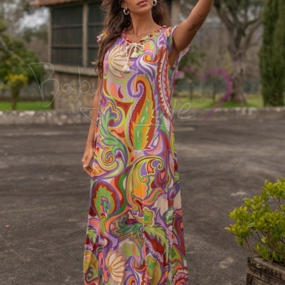 Vestido comprido colorido com padrão psicadélico e decote com laço, usado por mulher a tirar selfie ao ar livre.