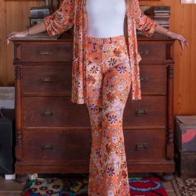 Conjunto de casaco e calças florais em tons de laranja com fundo branco, usado por mulher junto a móvel de madeira