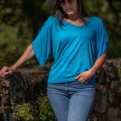 Mulher com túnica azul, chapéu de palha e óculos de sol junto a parede de pedra