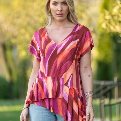 Blusa feminina com padrão abstrato em vermelho, rosa e laranja, decote em V, mangas curtas, bainha assimétrica, vista ao ar livre.