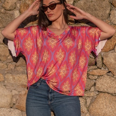 Mulher com t-shirt colorida com padrão floral e geométrico e jeans, óculos escuros, parede de pedra ao fundo
