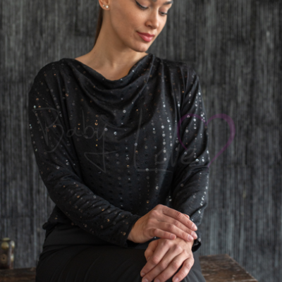 Blusa preta com pontos brilhantes e decote drapeado usada por mulher sentada em madeira escura