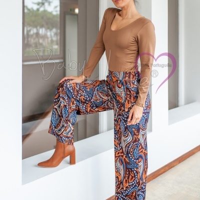 Mulher com body castanho e calças largas coloridas, botas castanhas, em ambiente interior branco