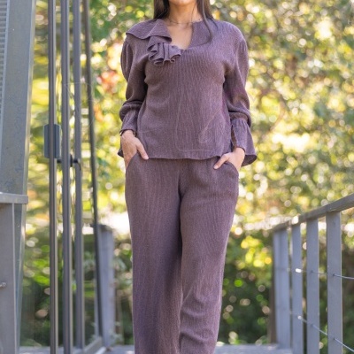 Mulher com conjunto castanho canelado composto por túnica com folho e calças largas, óculos escuros e botas castanhas, em ponte metálica com fundo de árvores.