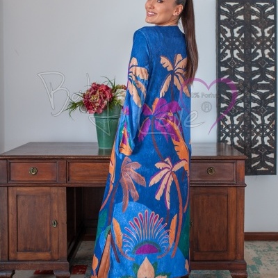 Vestido comprido azul com padrão de palmeiras e flores, botas castanhas, ambiente interior com mesa e decoração.