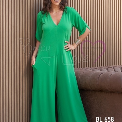 Mulher a usar macacão verde de tecido fluido com decote em V e mangas 3/4 em frente a parede de ripas de madeira e sofá castanho