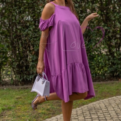 Mulher de vestido lilás solto com ombros descobertos, sandálias e mala lilás, em exterior com sebes ao fundo.