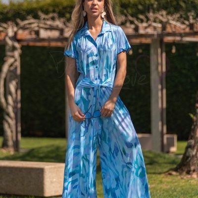 Mulher usando macacão azul largo com padrão abstrato em várias tonalidades de azul, botões frontais, cinto na cintura, ao ar livre
