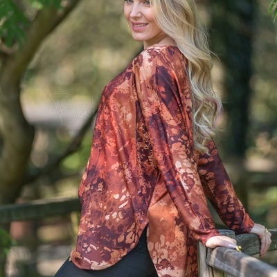 Mulher com túnica laranja castanha com padrão abstrato junto a corrimão de madeira em ambiente natural.