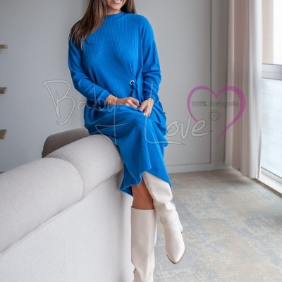 Mulher com vestido azul e botas brancas sentada em sofá cinzento em sala com cortinas claras