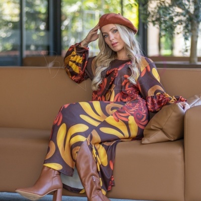 Mulher com vestido colorido, boina castanha e botas sentada num sofá castanho