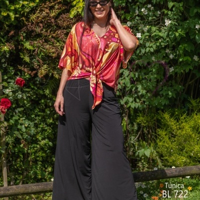Mulher de túnica colorida e pantalona preta ao ar livre com vegetação ao fundo