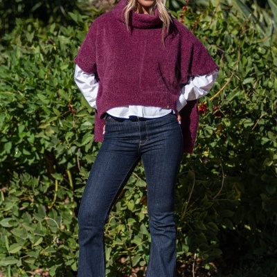 Mulher com capa bordeaux de malha, camisa branca, jeans e sapatos bordeaux, ao ar livre com plantas ao fundo