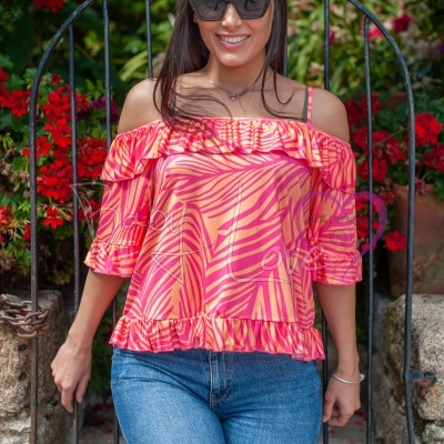 Mulher com blusa rosa laranja com padrão abstrato, mangas 3/4, babados, calças de ganga azuis, à frente de uma porta metálica e plantas