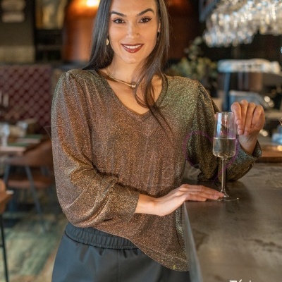 Mulher jovem com túnica dourada brilhante e saia preta segurando copo de champanhe num bar.