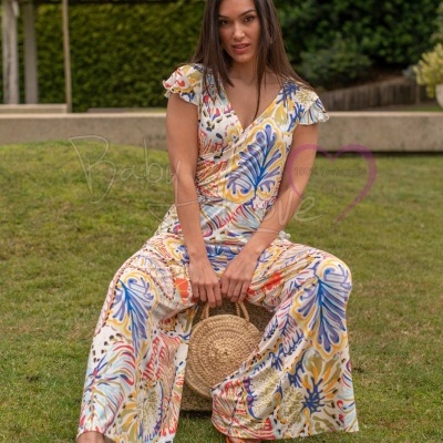 Mulher sentada numa relva a usar macacão estampado colorido e sandálias laranja, com bolsa de palha redonda.
