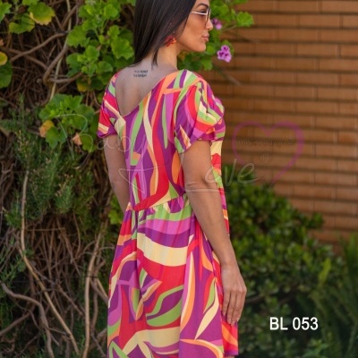 Vestido curto colorido com padrão abstrato em ambiente exterior com plantas e parede de tijolos.