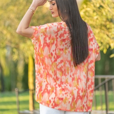 Blusa leve com padrão coral, rosa e amarelo em mulher com cabelo comprido, pulseira dourada, fundo de jardim