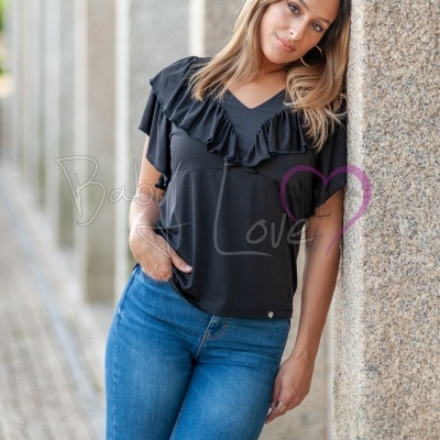 Mulher com blusa preta de folhos e calças de ganga azuis encostada a parede de pedra