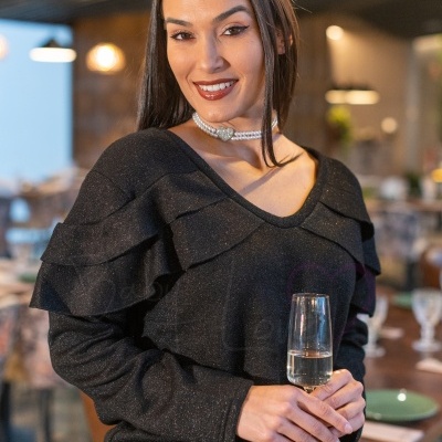 Mulher com túnica preta brilhante e folhos, colar de pérolas, segurando taça de vidro num restaurante