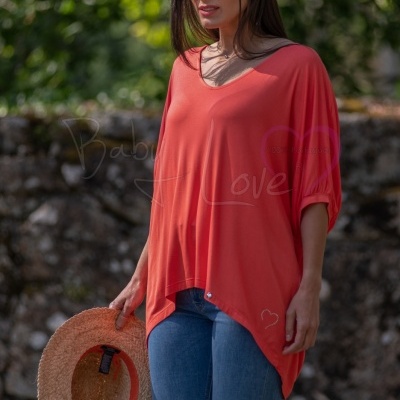 Mulher com túnica laranja e calças de ganga segurando chapéu de palha em ambiente exterior