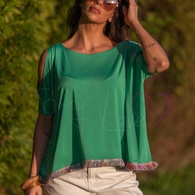 Mulher com blusa verde de tecido leve e calças brancas em ambiente exterior com vegetação.