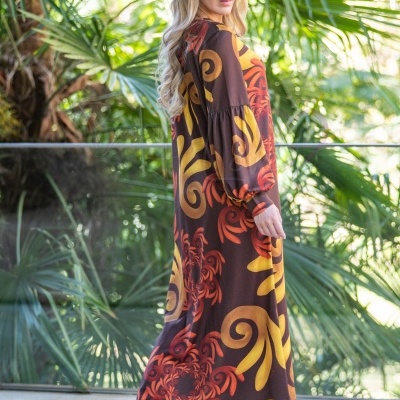 Mulher de perfil com vestido comprido colorido, botas castanhas e boina castanha no exterior com plantas verdes no fundo