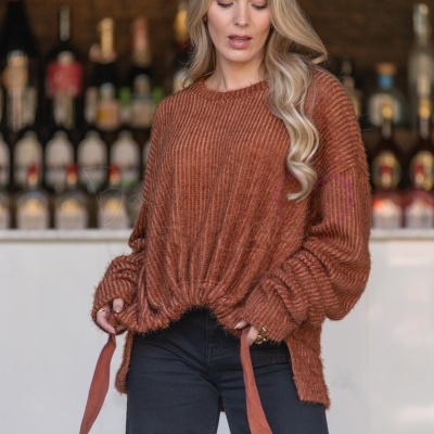 Mulher com camisola de malha castanha e calças pretas em frente a um bar com garrafas desfocadas
