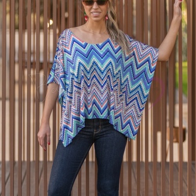 Mulher com blusa larga colorida com padrão zig-zag e jeans escuros em frente a uma vedação de madeira