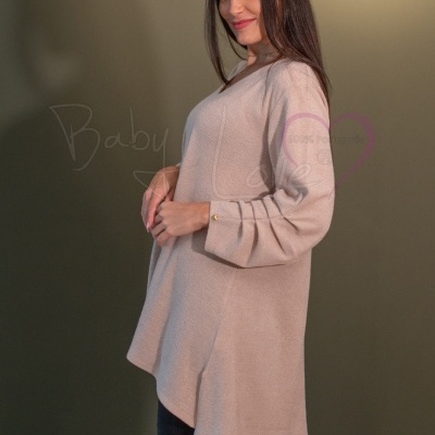 Mulher com túnica bege clara, calças pretas e botas castanhas frente a fundo verde-escuro