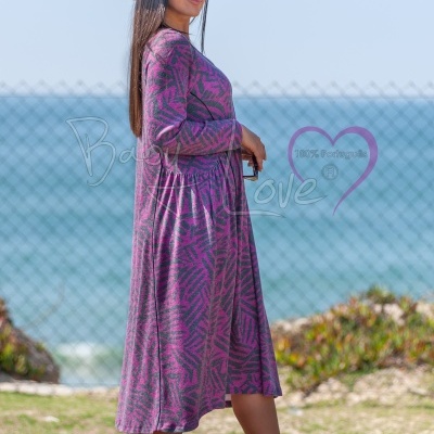 Mulher com vestido rosa e roxo padrão geométrico, botas pretas e chapéu preto numa paisagem ao ar livre com mar ao fundo