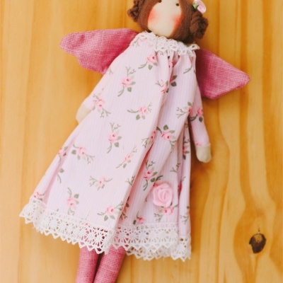 Boneca de pano com vestido rosa floral e asas cor-de-rosa sobre madeira