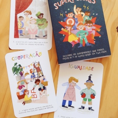 Caixa e cartas de jogo infantil com temas de superpoderes e valores como otimismo, cooperação e igualdade.