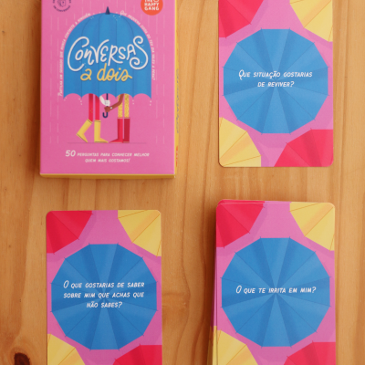 Jogo de cartas Conversas a Dois com caixa rosa e cartas coloridas sobre mesa de madeira
