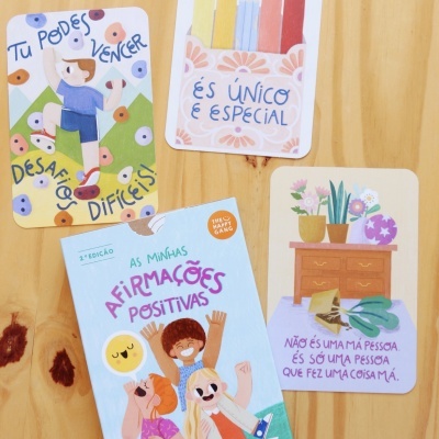Livro infantil com afirmações positivas e cartões coloridos com mensagens motivacionais em português sobre mesa de madeira clara