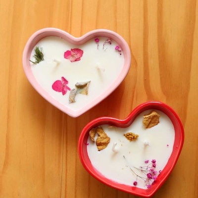 Velas em forma de coração, decoradas com flores secas, em bases rosa e vermelha sobre madeira clara