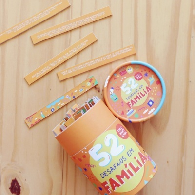 Caixa cilíndrica aberta laranja com tiras de papel e texto '52 Desafios em Familia' sobre madeira clara