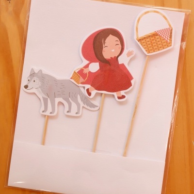 Três palitos decorativos com personagens de papel, incluindo um lobo, menina com capuz vermelho e uma cesta, sobre base branca e fundo de madeira.