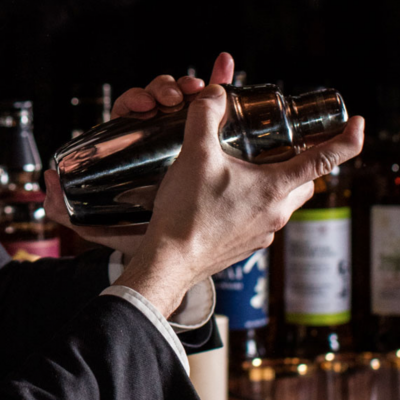 Coqueteleira metálica em mãos com garrafas ao fundo