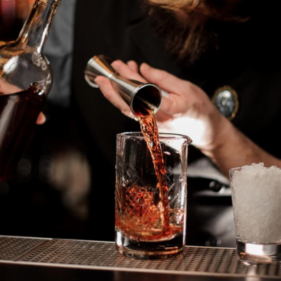 Pessoa a servir bebida castanha num copo de vidro com um medidor metálico ao lado de copo com gelo picado.