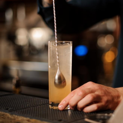 Copo alto com bebida clara e gelo sendo mexida com colher de bar em ambiente de bar