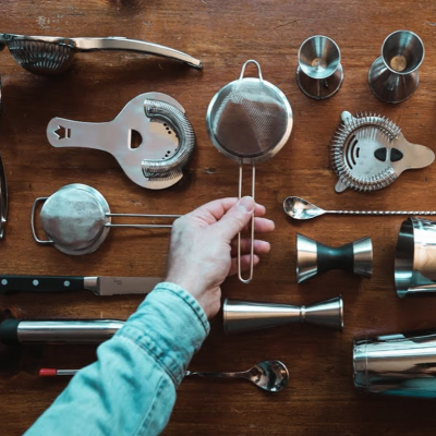 Utensílios de bar metálicos prateados sobre mesa de madeira com mão segurando peneira