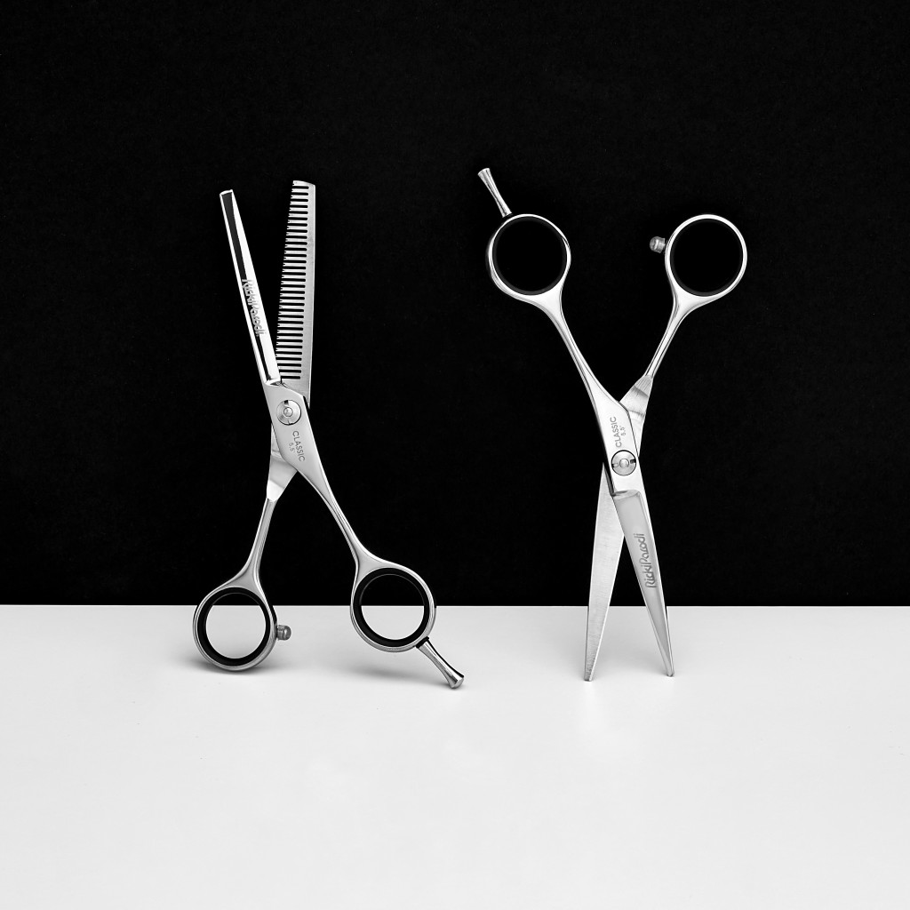 Duas tesouras de cabelo em aço inoxidável, uma lisa e uma dentada, sobre fundo preto e superfície branca