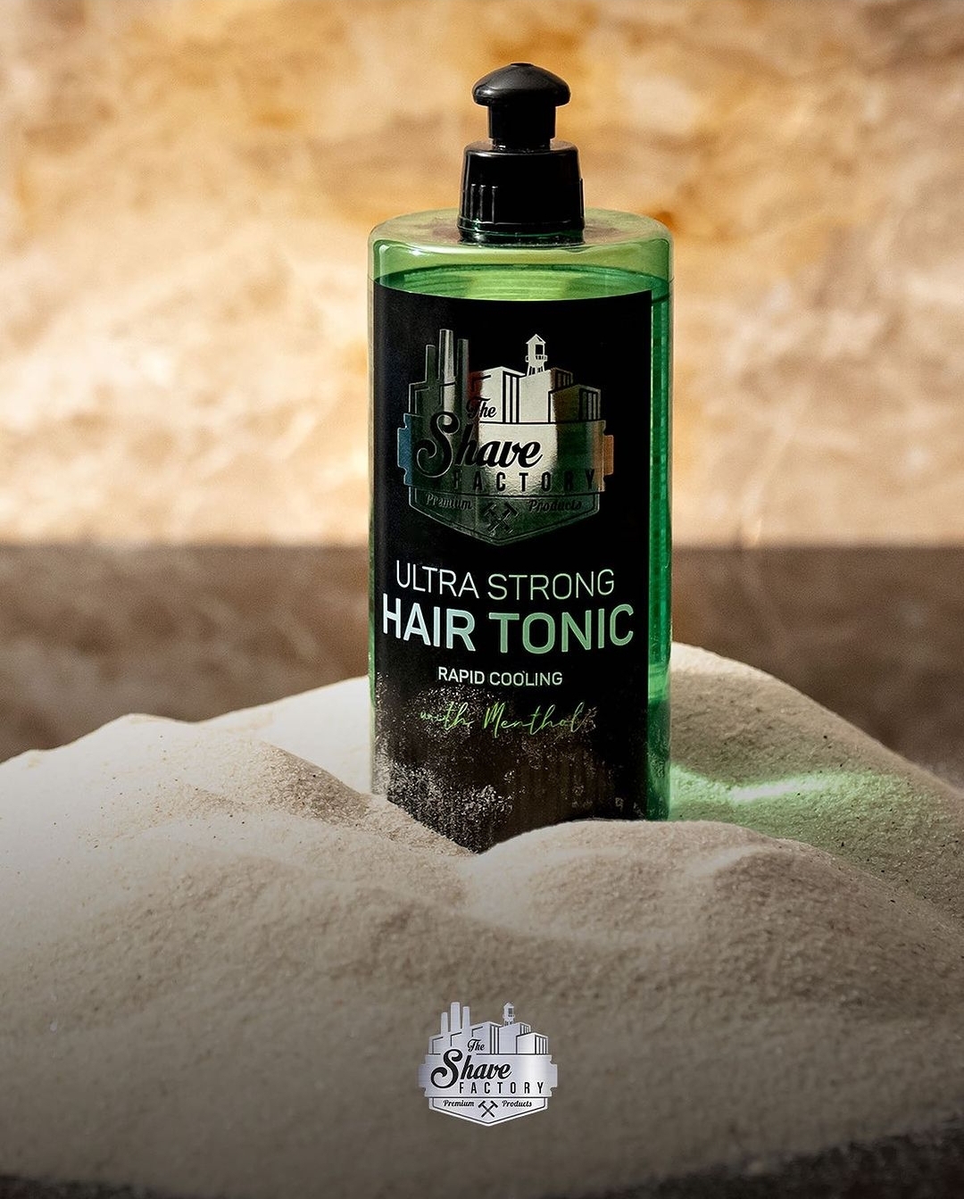 Frasco verde com tónico capilar sobre areia branca, rótulo preto com texto The Shave Factory Ultra Strong Hair Tonic