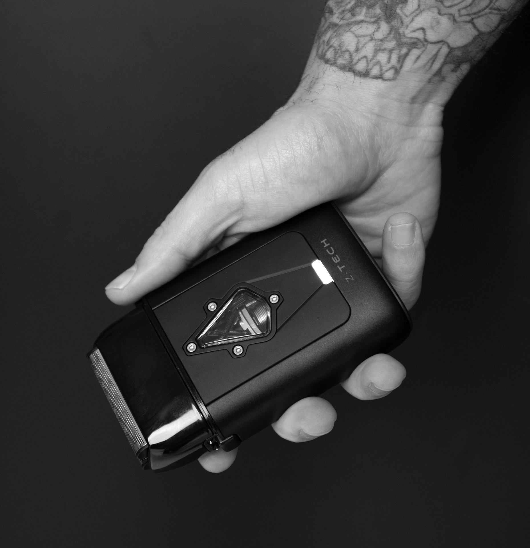 Mão tatuada a segurar dispositivo eletrônico preto com luz e texto ZTECH em fundo escuro