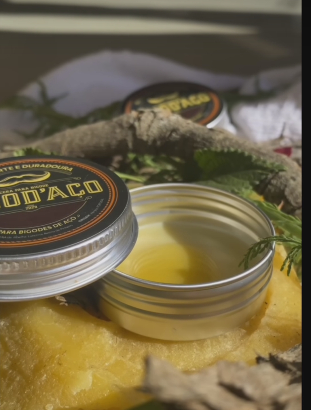 Pote aberto com cera para bigodes e tampa preta com texto amarelo, rodeado de folhas verdes e textura amarela